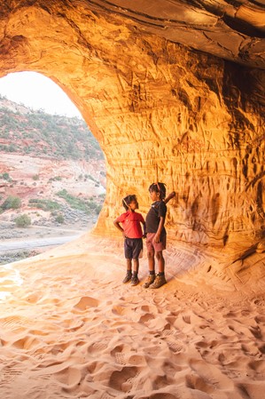 Two children standing in a sandy cave illuminated by sunlight.