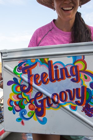 A woman smiling and holding a handwritten sign that reads 'feeling groovy,' with a raft visible behind her at a lake.