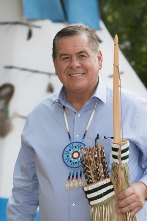 A man wearing a blue button-down shirt stands in front of a white canvas teepee, holding culturally significant artifacts.