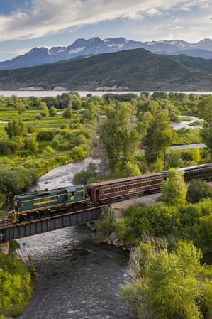 A train crossing a bridge over a stream in a valley, with mountains in the background.
